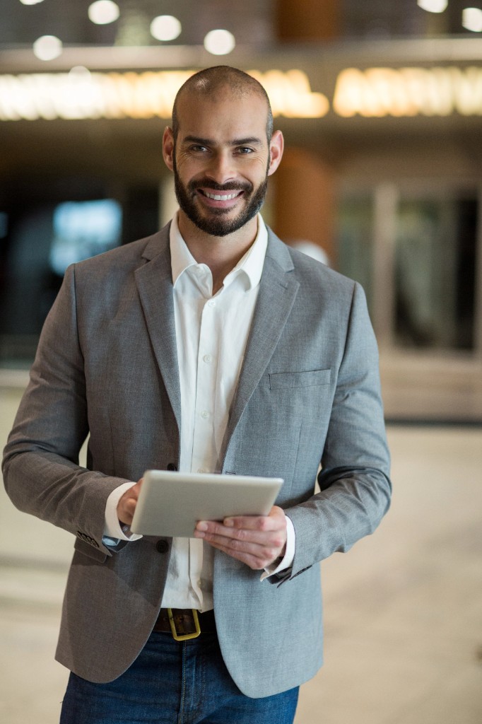 A smiling man in a grey blazer holds a tablet in a lobby